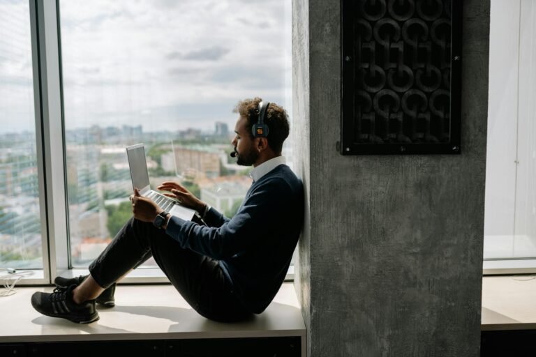 A man using a laptop while sitting by a window in an office, wearing headphones.
