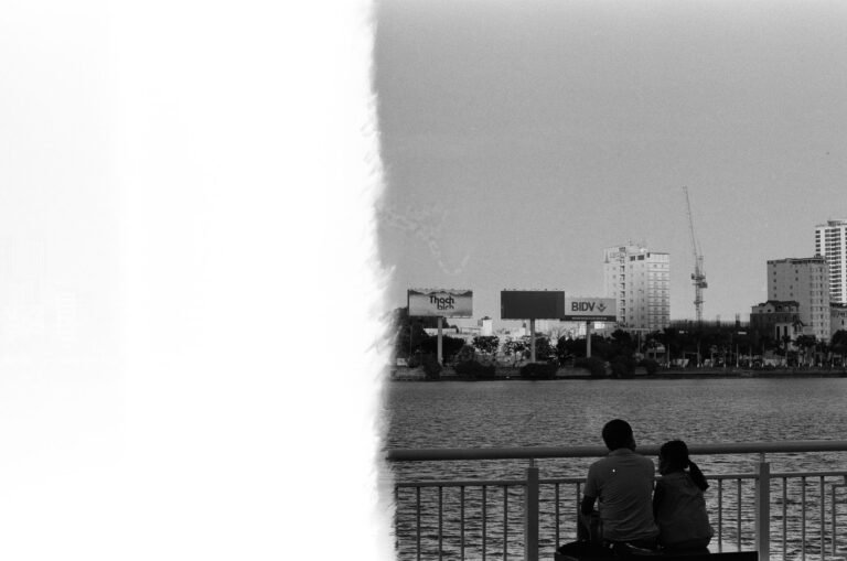 Monochrome image of a couple sitting by the Đà Nẵng river, viewing the city skyline.