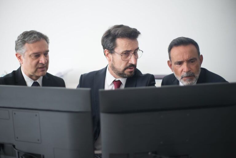Team of three businessmen in suits and glasses collaborating at desks, focused on shared screens.